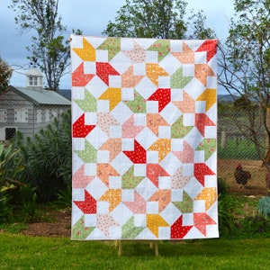 May include: A white quilt with a patchwork design featuring red, yellow, pink, and green fabric squares. The quilt is hanging on a clothesline in a backyard setting.
