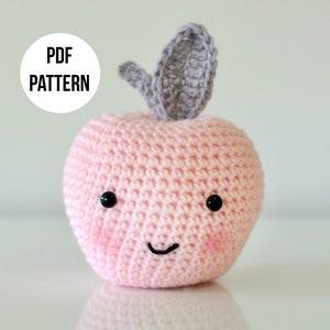 May include: A pink crocheted apple with a grey crocheted leaf on top. The apple has a smiling face with black eyes and a black mouth.