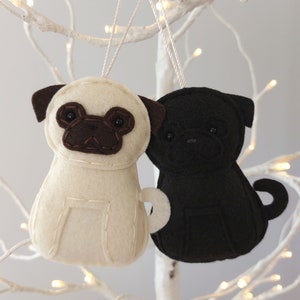 May include: Two felt pug dog ornaments, one white and one black, hanging from a white branch. Both pugs have brown felt noses and eyes.