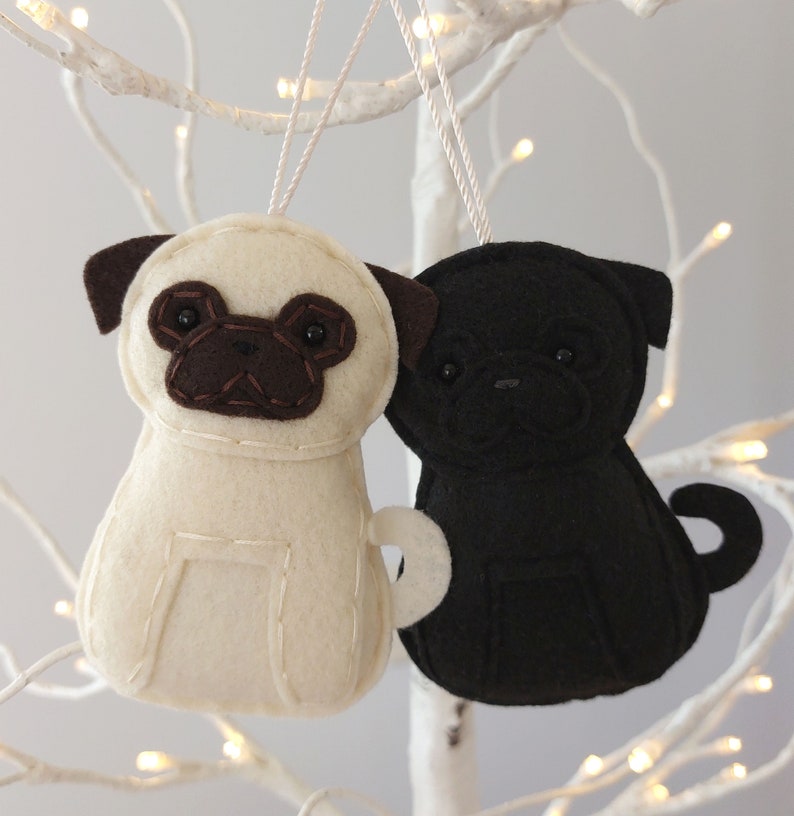 May include: Two felt pug dog ornaments, one white and one black, hanging from a white branch. Both pugs have brown felt noses and eyes.