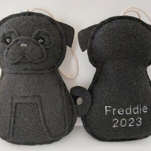 May include: Black felt pug ornament with a white string for hanging. The back of the ornament has the name "Freddie" and the year "2023" embroidered in white.