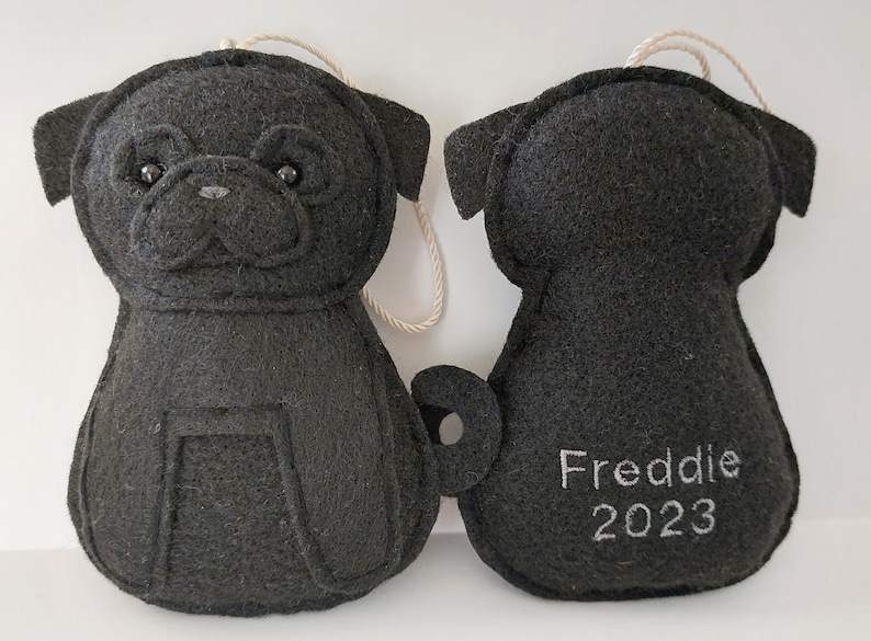 May include: Black felt pug ornament with a white string for hanging. The back of the ornament has the name "Freddie" and the year "2023" embroidered in white.