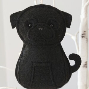 May include: Black felt pug dog ornament hanging from a white branch. The ornament has black felt eyes and a black felt nose.