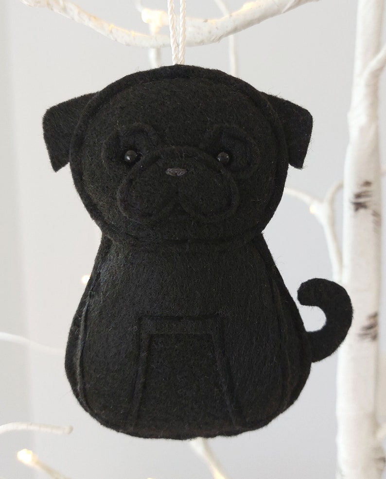 May include: Black felt pug dog ornament hanging from a white branch. The ornament has black felt eyes and a black felt nose.