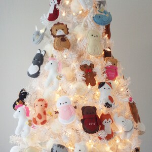 May include: A white artificial Christmas tree decorated with felt ornaments. The ornaments are shaped like animals, including a unicorn, a giraffe, a bear, a fox, a cat, a penguin, a whale, a llama, a snowman, a gingerbread man, and a bird. Some of the ornaments have personalized names and dates, such as "Emma 2019" and "Charlie 2019".