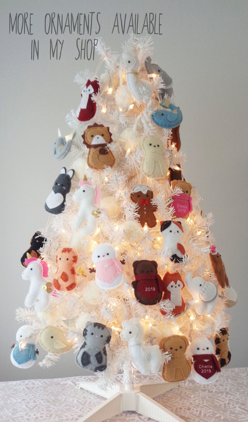 May include: A white artificial Christmas tree decorated with felt ornaments. The ornaments are shaped like animals, including a unicorn, a giraffe, a bear, a fox, a cat, a penguin, a whale, a llama, a snowman, a gingerbread man, and a bird. Some of the ornaments have personalized names and dates, such as "Emma 2019" and "Charlie 2019".