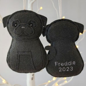 May include: Black felt Christmas ornament shaped like a pug dog. The ornament has a stitched face and the text "Freddie 2023" on the back.