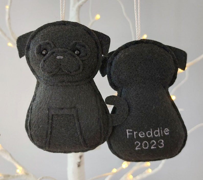 May include: Black felt Christmas ornament shaped like a pug dog. The ornament has a stitched face and the text "Freddie 2023" on the back.