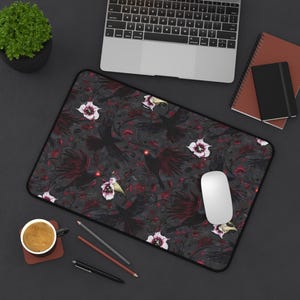 Puede incluir: Alfombrilla de ratón rectangular con un fondo gris oscuro que presenta un patrón de cuervos negros con ojos rojos, flores blancas y detalles en rojo. Un ratón de ordenador blanco está sobre la alfombrilla, junto a un portátil.
