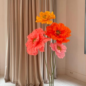May include: A set of four large paper flowers in shades of orange, pink, and red. The flowers are arranged on a black stand and are displayed against a beige curtain.