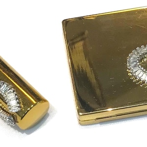 May include: A gold-toned cylindrical lipstick case and a matching square compact mirror, both featuring a horseshoe-shaped embellishment encrusted with clear baguette-cut crystals on their polished, reflective metallic surfaces.