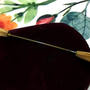 May include: A gold-toned metal lapel pin featuring a long, thin central stem connecting two elongated, ribbed, spindle-shaped ends. Each end is capped with a small, round, white bead. The pin is positioned horizontally across a dark maroon velvet surface.