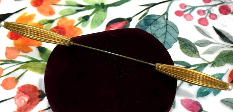 May include: A gold-toned metal lapel pin featuring a long, thin central stem connecting two elongated, ribbed, spindle-shaped ends. Each end is capped with a small, round, white bead. The pin is positioned horizontally across a dark maroon velvet surface.