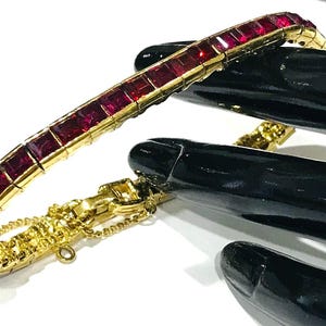 Pode incluir: Uma pulseira dourada com uma fileira de pedras preciosas retangulares vermelho-escuras. A pulseira é mostrada em um fundo branco, com objetos pretos e brilhantes ao fundo. A pulseira tem uma corrente e fecho delicados.
