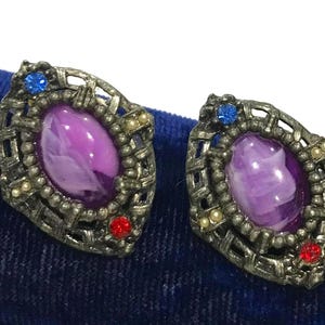 May include: A pair of silver-toned metal earrings featuring an oval-shaped purple marbled cabochon center. The frame has a textured lattice design accented with a single small blue rhinestone at the top and a single small red rhinestone at the bottom.