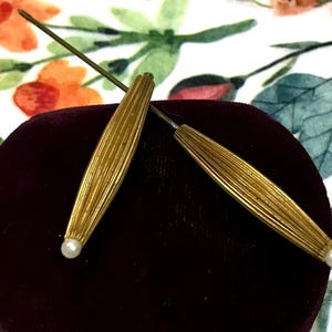 May include: Two gold-toned metal hat pins featuring elongated, ribbed, spindle-shaped heads. Each pin is finished with a small, round, white bead at the pointed end. The pins are positioned in a crossed arrangement, showcasing their metallic texture.