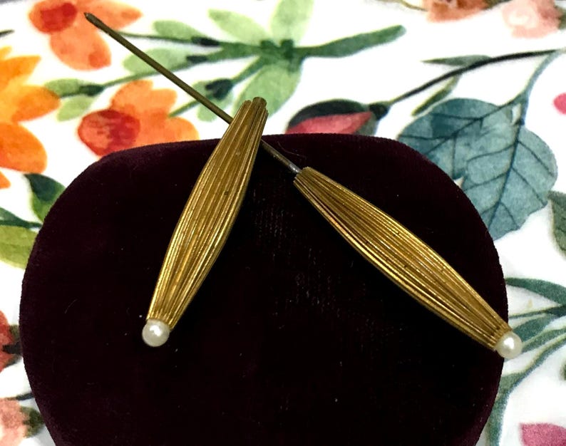 May include: Two gold-toned metal hat pins featuring elongated, ribbed, spindle-shaped heads. Each pin is finished with a small, round, white bead at the pointed end. The pins are positioned in a crossed arrangement, showcasing their metallic texture.