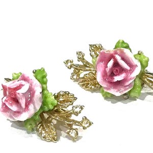 Puede incluir: Un par de pendientes dorados con una rosa central de cer&aacute;mica rosa y blanca rodeada de hojas de cer&aacute;mica verde y peque&ntilde;os brillantes transparentes. La base met&aacute;lica presenta un dise&ntilde;o de hojas texturizadas en cada pieza.