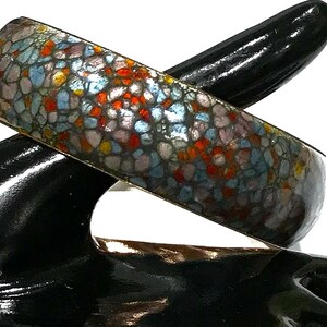 May include: A mosaic-patterned bangle bracelet. The bracelet displays a pattern of small, irregular shapes in red, blue, and grey. The bracelet is presented on a black, glossy surface.