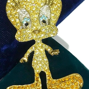 May include: A brooch shaped like the cartoon character Tweety Bird, covered in yellow and clear crystals. The head and body feature light yellow stones, while the large feet are set with darker orange-yellow crystals. The eyes have white stones.