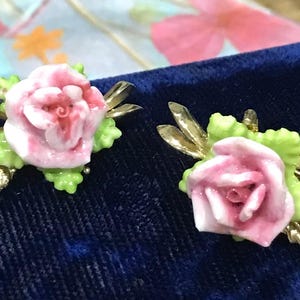 Puede incluir: Un par de pendientes con rosas de cer&aacute;mica rosa esculpidas y hojas de color verde claro, sobre detalles de metal dorado. Cada pendiente incluye peque&ntilde;os diamantes de imitaci&oacute;n transparentes incrustados en el dise&ntilde;o de hojas met&aacute;licas.