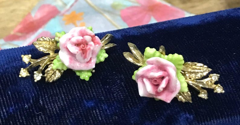 Puede incluir: Un par de pendientes con rosas de cer&aacute;mica rosa esculpidas y hojas de color verde claro, sobre detalles de metal dorado. Cada pendiente incluye peque&ntilde;os diamantes de imitaci&oacute;n transparentes incrustados en el dise&ntilde;o de hojas met&aacute;licas.