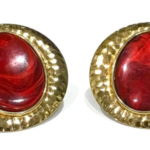 May include: A pair of vintage clip-on earrings. Each earring features a large, oval, red stone with a marbled appearance, set in a hammered gold-tone metal frame. The earrings are a bold statement piece.