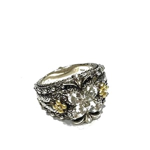 May include: A sterling silver ring featuring a central four-petal flower motif set with clear stones, accented by smaller gold-toned floral details. The band has a textured, oxidized finish with intricate metalwork and is marked 18K and 925.