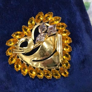 May include: A heart-shaped gold-tone brooch featuring a border of marquise-cut amber-colored crystals. The center includes a smooth, polished gold-tone metal swirl design accented with three small, light purple diamond-shaped crystals at the top.