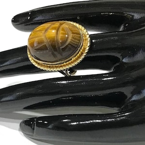 May include: A gold ring featuring an oval-shaped, brown and gold tiger's eye gemstone carved with a scarab beetle design. The stone is set in a gold-toned metal bezel with a twisted rope-style border detail around the edge of the setting.