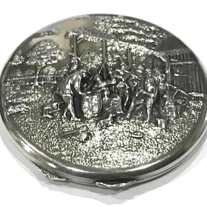 May include: A silver compact with a raised design of people in a village setting. The compact has a hinged lid and a smooth, reflective surface.