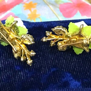 Puede incluir: Dos broches de metal en tono dorado con dise&ntilde;os florales blancos y hojas verdes. El reverso de cada broche muestra un mecanismo de alfiler con cierre giratorio, dejando ver la estructura met&aacute;lica texturizada y la parte trasera de los p&eacute;talos.