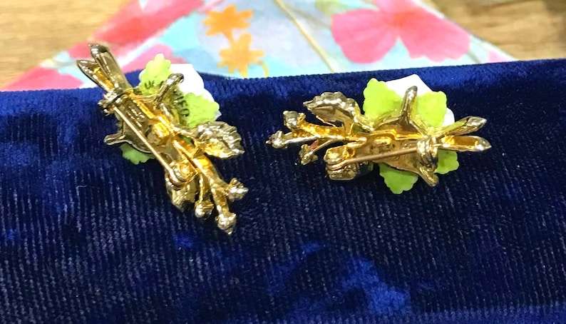 Puede incluir: Dos broches de metal en tono dorado con dise&ntilde;os florales blancos y hojas verdes. El reverso de cada broche muestra un mecanismo de alfiler con cierre giratorio, dejando ver la estructura met&aacute;lica texturizada y la parte trasera de los p&eacute;talos.