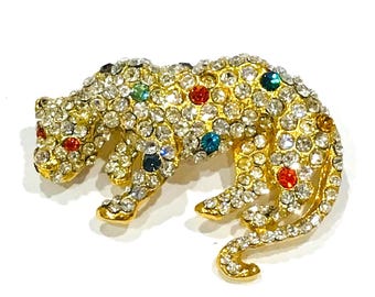 May include: A gold-toned brooch in the shape of a leopard, encrusted with clear and colorful rhinestones. The leopard is depicted in a crouching position, with its tail curled. The rhinestones are in various colors, including red, blue, and green.