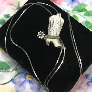 May include: A silver-toned necklace featuring a cowboy boot pendant with an attached spur. The boot has intricate engraved detailing on the shaft and a row of small clear stones along the ankle. The pendant hangs from a thin, reflective silver-toned chain.