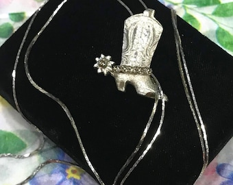 Op de afbeelding: Een zilverkleurige ketting met een cowboylaars-hanger inclusief spoor. De laars heeft gedetailleerde gravures op de schacht en een rij kleine, heldere steentjes bij de enkel. De hanger hangt aan een dunne, reflecterende zilverkleurige ketting.