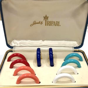 May include: A vintage jewelry box with the Trifari logo contains a pair of blue hoop earrings and several pairs of colorful, curved earrings in red, coral, turquoise, and white. The box is blue with a white interior.