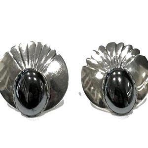 May include: A pair of circular earrings featuring a polished silver-tone metal base with a fan-like pleated design at the top. Each earring is centered with a smooth, oval-shaped black stone that has a reflective, glossy finish and a dark appearance.