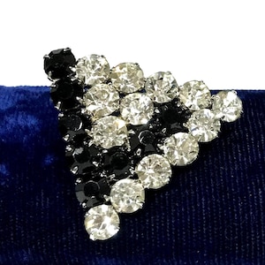 May include: A triangular brooch featuring a geometric arrangement of round, faceted clear and black rhinestones set in metal. The design consists of alternating rows and clusters of dark black stones and sparkling clear stones in a pointed shape.