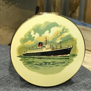 May include: A round, vintage compact with a cream-colored background. The compact features a detailed illustration of the RMS Ivernia cruise ship sailing on green water, with a sky of green and yellow clouds. The ship is black and white with red accents.