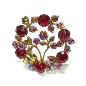 May include: A vintage gold-tone brooch with a floral design. The brooch features large, round, red gemstones and smaller iridescent stones. The design includes stylized leaves and stems, creating a circular arrangement.