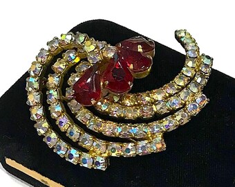 Pode incluir: Um broche de tom dourado com três pedras vermelhas proeminentes em forma de lágrima no centro, rodeadas por várias filas curvas de pequenos strass transparentes e iridescentes que criam um design em leque sobre a estrutura metálica.
