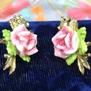 Puede incluir: Un par de pendientes que presentan una rosa de cer&aacute;mica rosa y blanca en el centro con hojas de color verde brillante, acentuadas por elementos de follaje de metal dorado. El dise&ntilde;o floral muestra p&eacute;talos y tallos detallados y delicados.