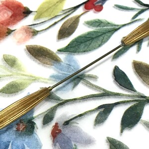 May include: A long, thin metal hat pin featuring two elongated, ribbed gold-tone metal caps at each end, each tipped with a small white bead. The pin has a slender, straight metal shaft connecting the two decorative ends.