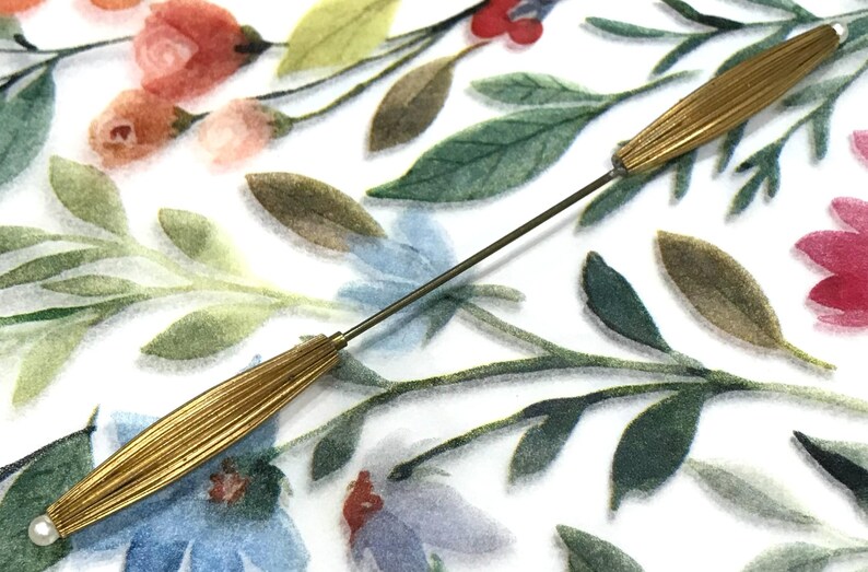 May include: A long, thin metal hat pin featuring two elongated, ribbed gold-tone metal caps at each end, each tipped with a small white bead. The pin has a slender, straight metal shaft connecting the two decorative ends.