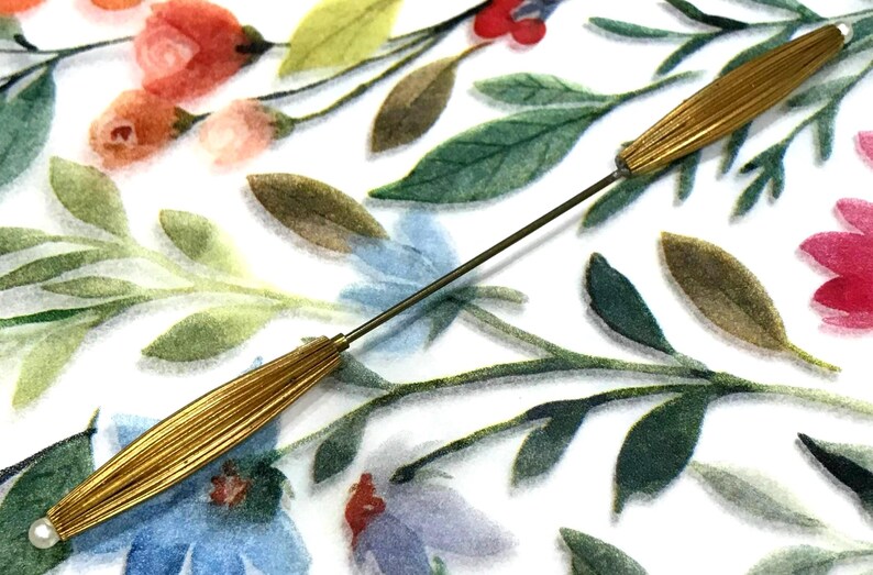May include: A long, thin metal hat pin featuring two elongated, ribbed gold-tone metal sections separated by a slender rod. Each end of the pin is capped with a small, round white bead, creating a symmetrical design for securing headwear.