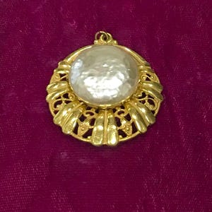 Puede incluir: Colgante dorado con una perla grande, redonda y blanquecina en el centro. El colgante tiene un diseño calado ornamentado con un borde festoneado. El fondo es de color magenta oscuro.