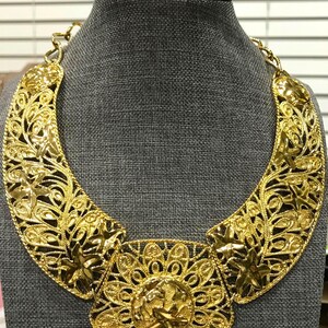 May include: A gold-coloured statement necklace with intricate filigree and floral patterns. The necklace features a central pendant with a circular design and a detailed figure. The necklace is displayed on a grey mannequin.