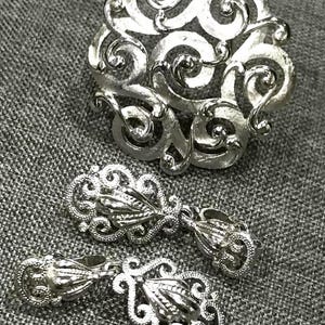 Pode incluir: Um conjunto de joias prateadas com um broche redondo com um design em espiral e dois brincos pendurados a condizer. Os brincos e o broche têm um estilo ornamentado e filigranado. O conjunto é exibido numa superfície cinzenta texturizada.