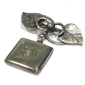 Peut inclure: Broche argentée avec un motif de feuilles et de volutes, et un pendentif carré avec un monogramme gravé. La broche présente deux feuilles et un élément décoratif en volute. Le pendentif est suspendu à une petite boucle.